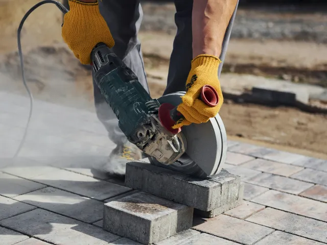 A pair of hands in work gloves hold a circular saw to cut a brick