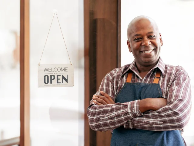 Man wearing Apron and checkered short smiling and standing in front of door with open sign to illustrate article about Small Business Saturday.