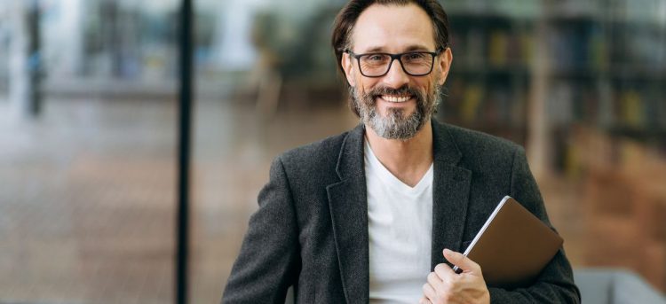 Portrait of happy confident businessman in eyaglasses and stylish formal suit. Middle aged successful male business leader looking directly at the camera, smiling, holding laptop in arms