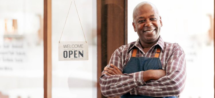 Portrait African barista or waiter cafe or coffee shop owner against entrance with open signboard, smiling guy in apron standing outdoors being proud of his small local business