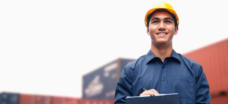 Smiling young engineer in protective work wear in a shipping yard examining cargo